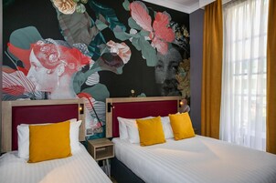 A hotel room with two beds, each with white and yellow pillows, and a large floral mural on the wall.