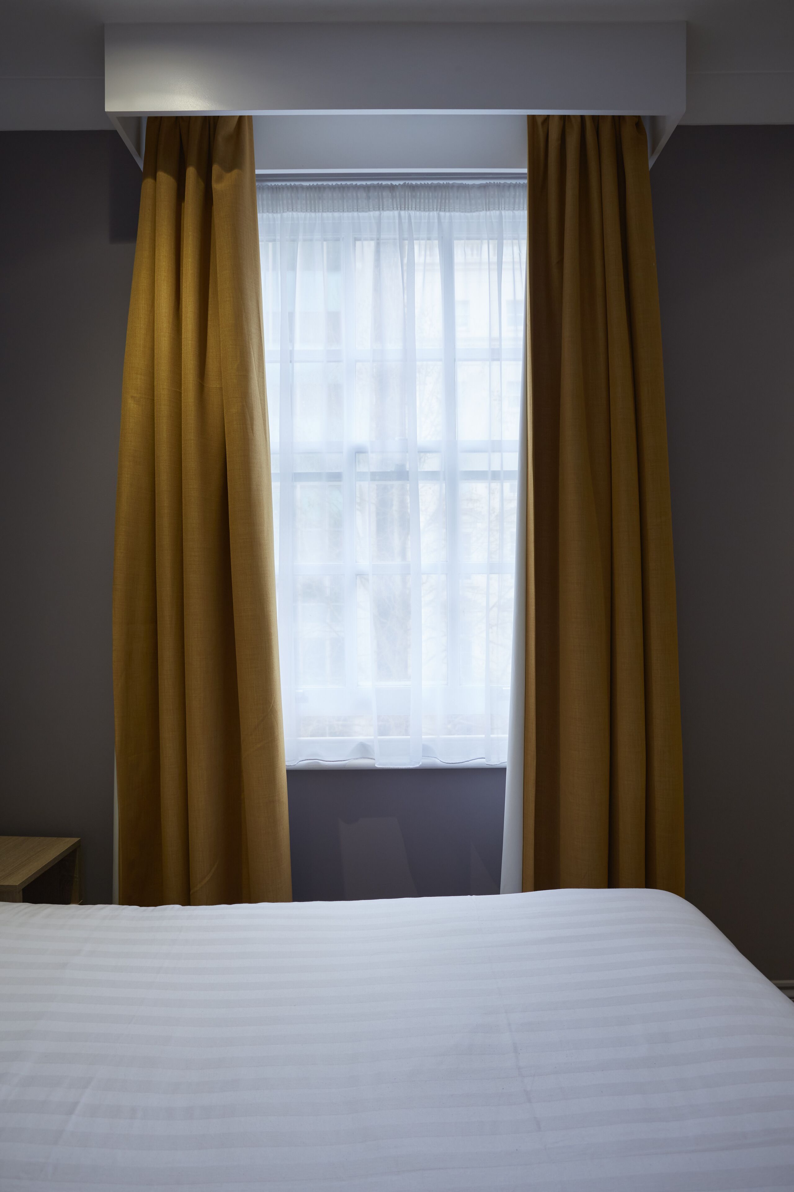Hotellihuone, jossa on suuri sänky, ikkuna, jossa on ohuet verhot, ja pelkistetty seinä.