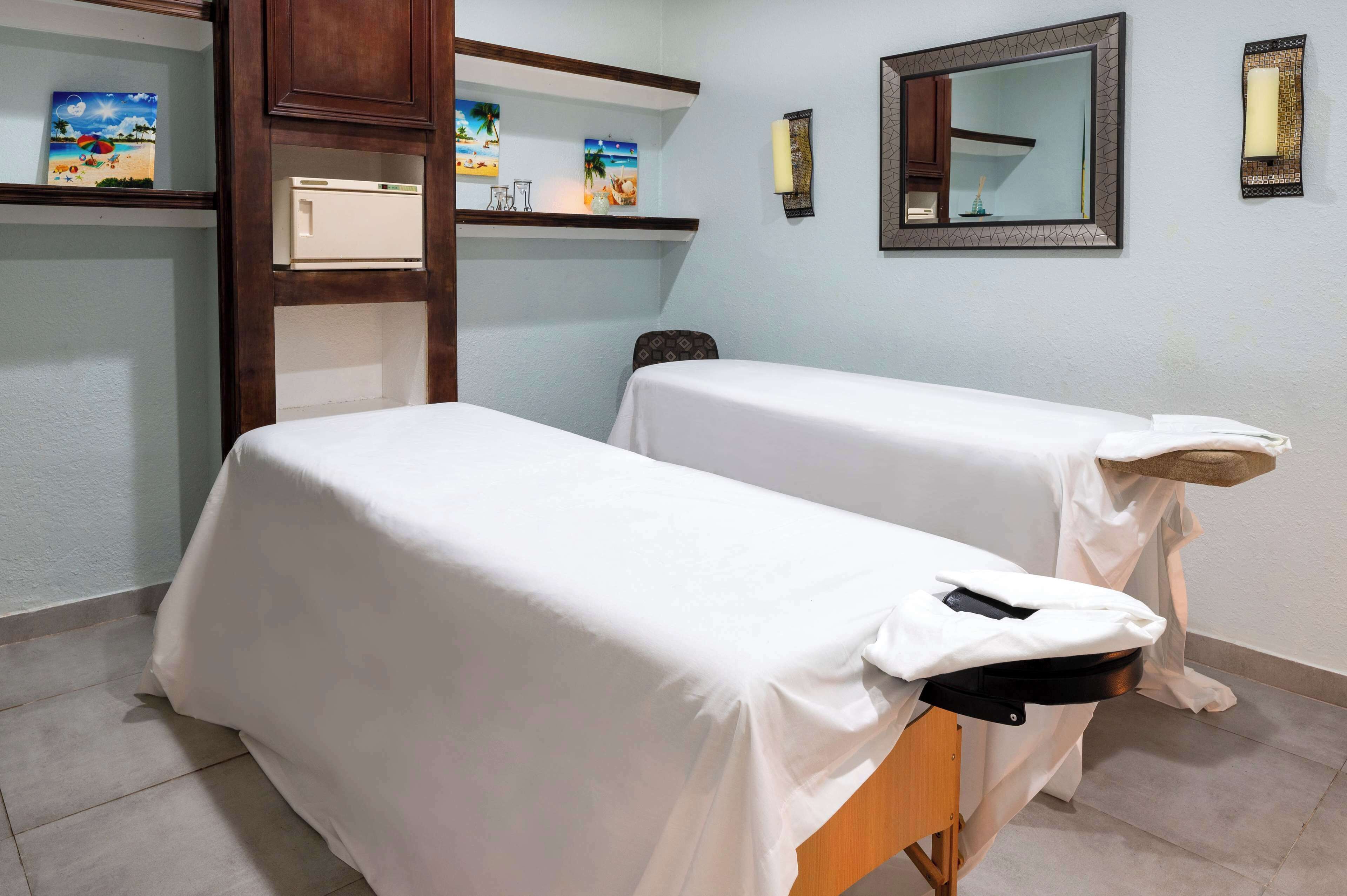 A room with two beds covered in white sheets, a wooden cabinet with artwork, a mirror, and a wall-mounted light fixture.
