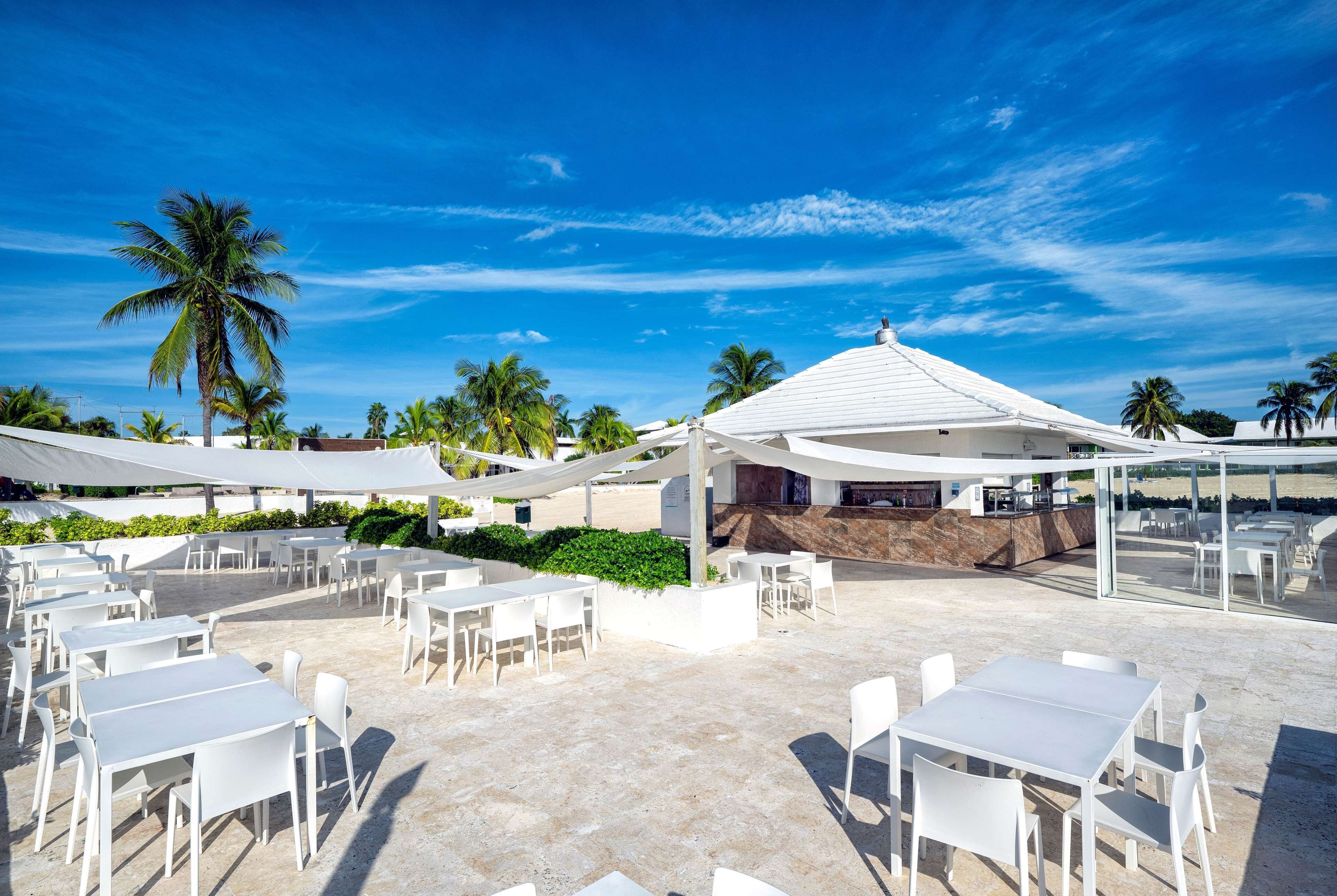 An outdoor patio area with white tables and chairs, palm trees, and a white building with a sloped roof.