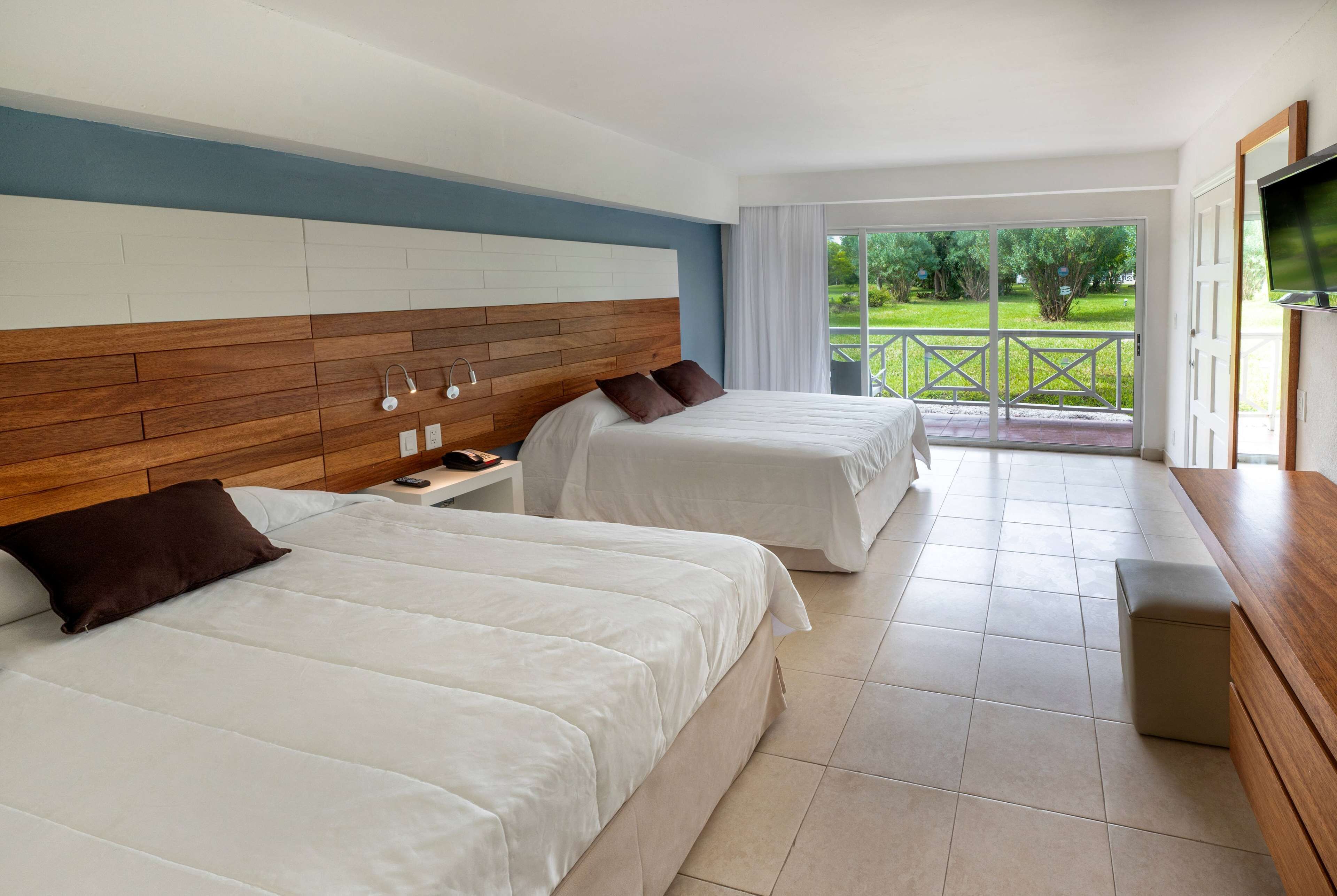 Ein Hotelzimmer mit zwei Betten, einem Holzkopfende, einem Fernseher und einem Balkon mit Blick auf Grünflächen.