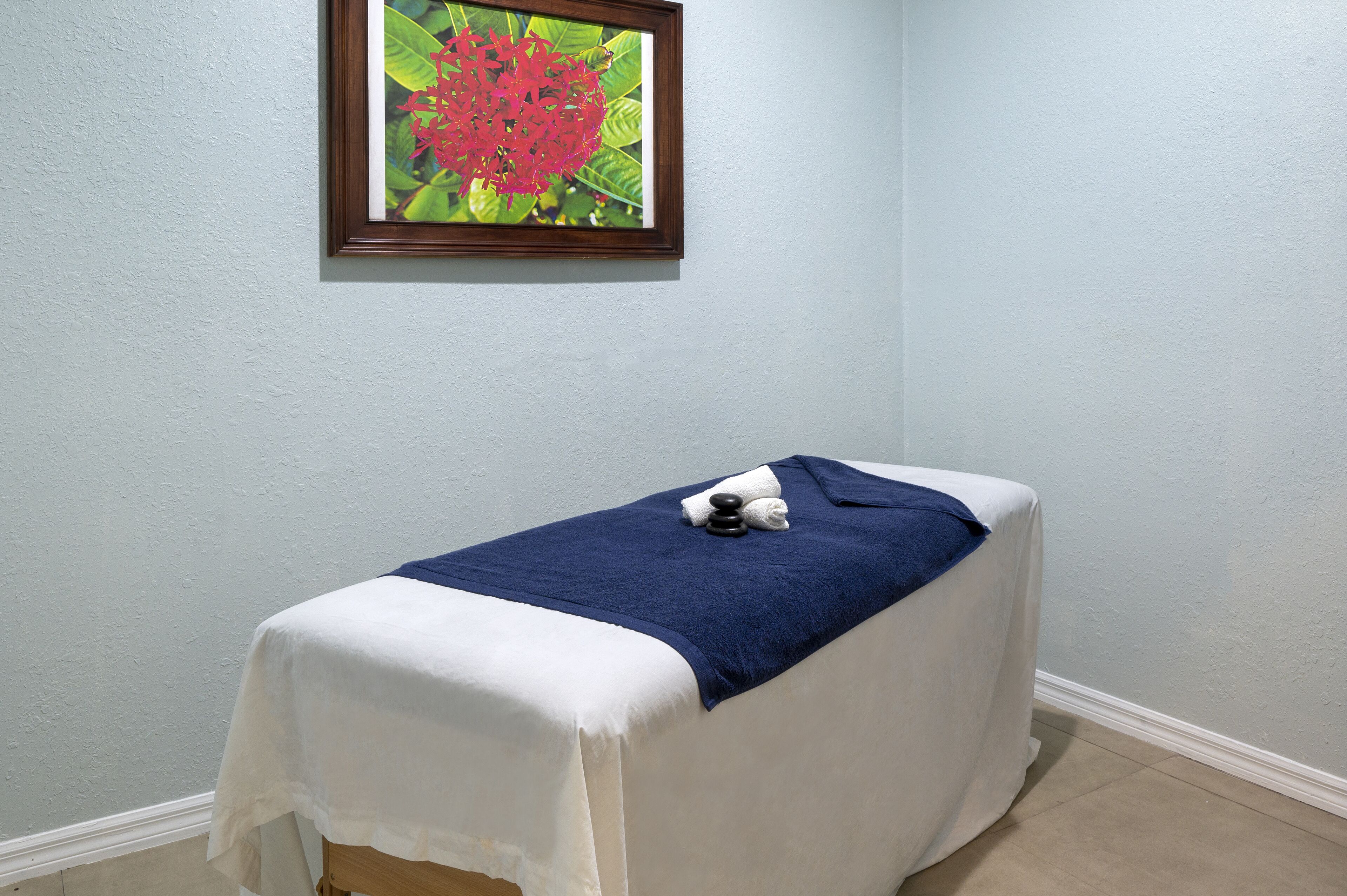 A single bed with a white bedspread and a blue towel, a framed picture of a flower on the wall, and a plain light blue wall.