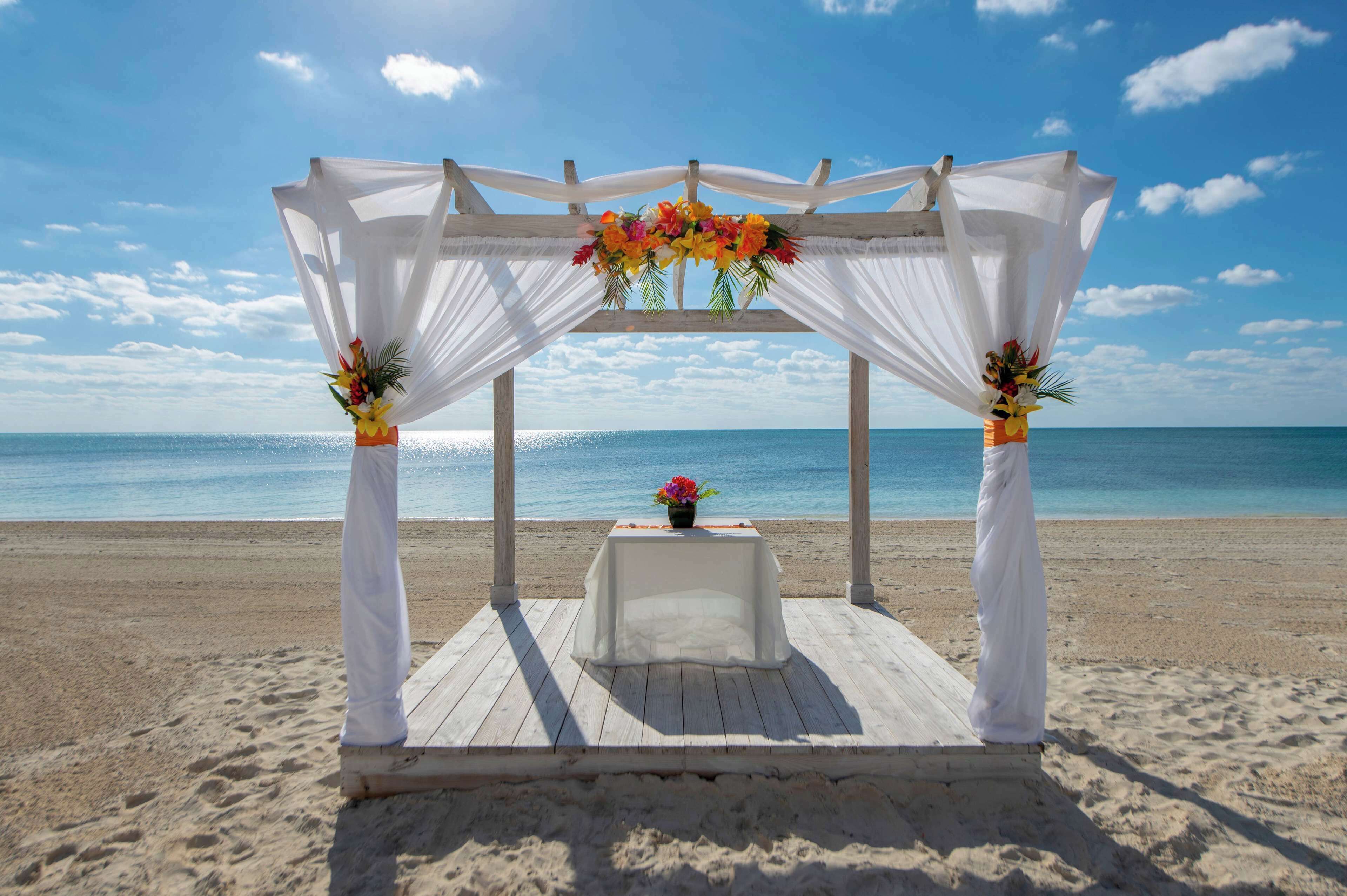 Montaje para una boda en la playa con un dosel blanco, decoración floral y una mesa.