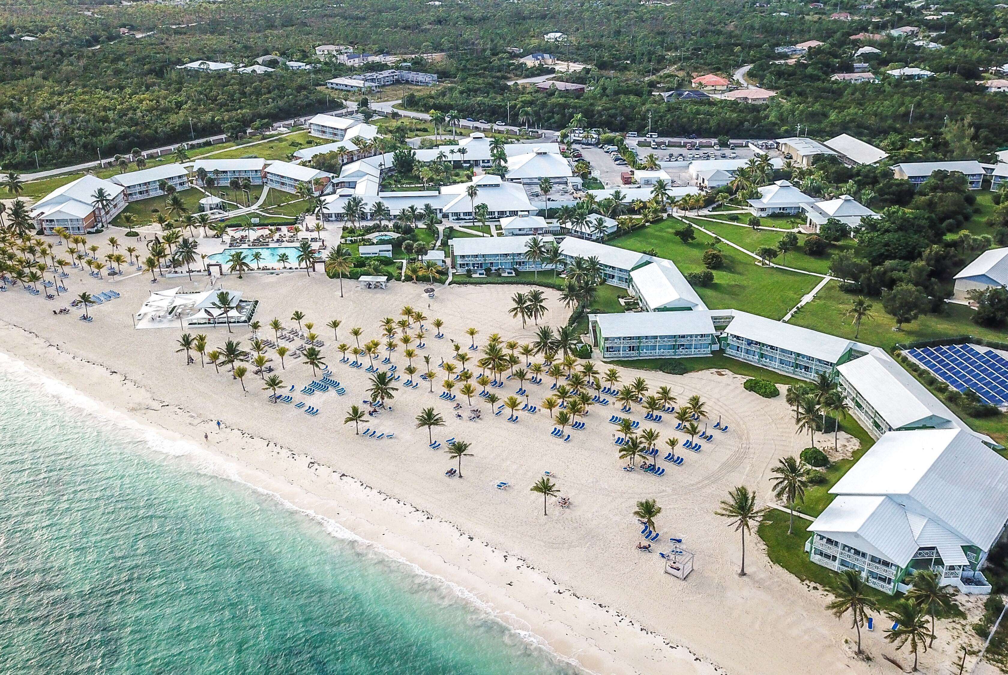 An aerial view of a resort with multiple buildings, a beach with palm trees and sun loungers, and a clear view of the ocean.