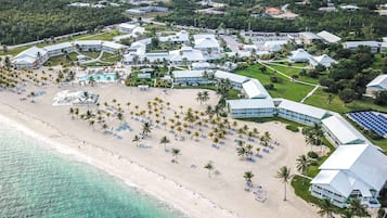 An aerial view of a resort with multiple buildings, a beach with palm trees and sun loungers, and a clear view of the ocean.