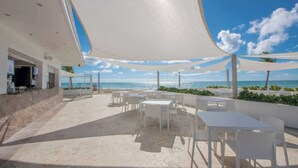 Un espace extérieur aménagé avec des tables et des chaises blanches, une vue sur la plage et une partie couverte avec un auvent blanc.