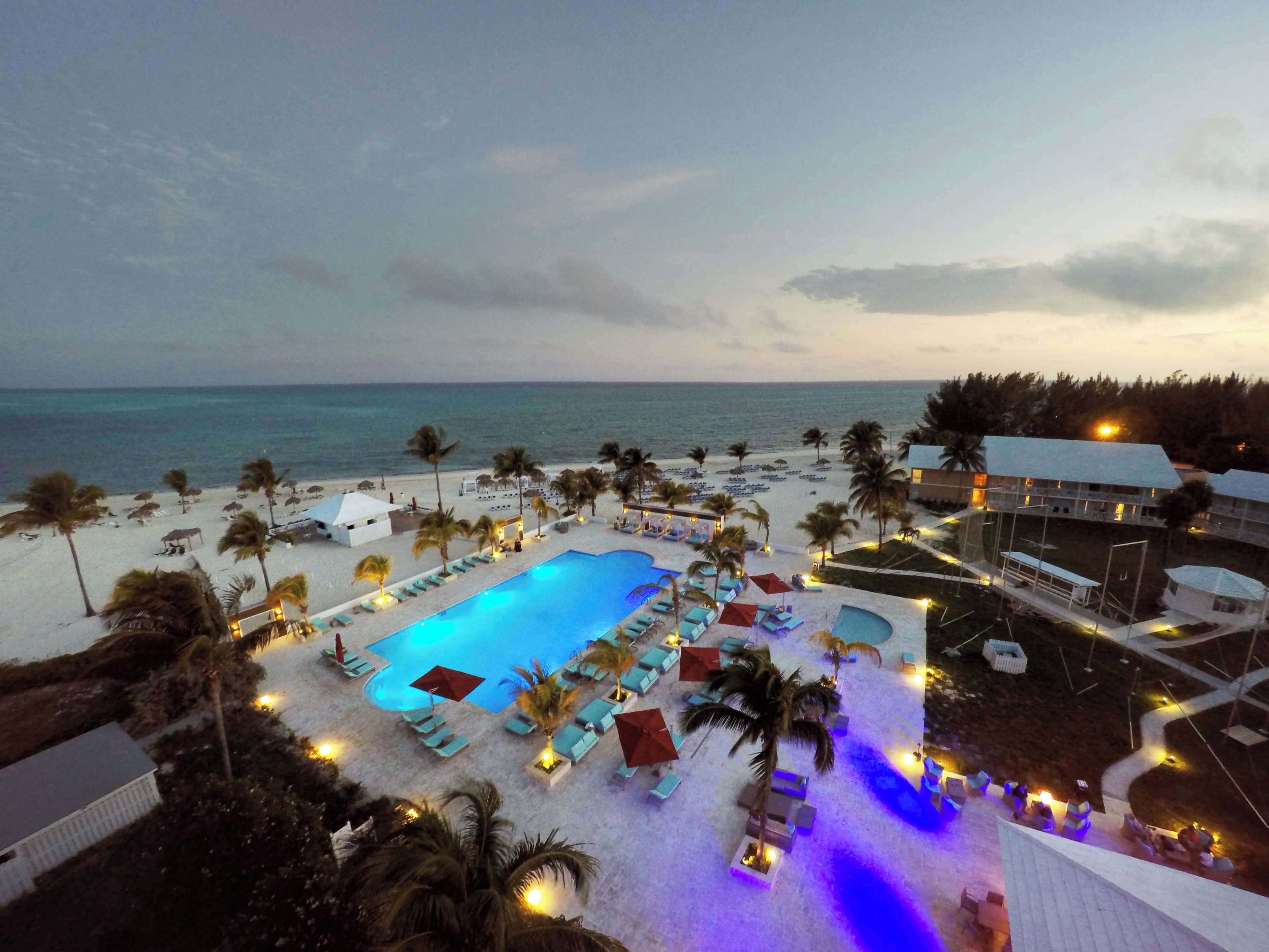 Um resort com uma piscina grande cercada por espreguiçadeiras e guarda-sóis, uma área de praia com guarda-sóis e espreguiçadeiras, e vista para o oceano.