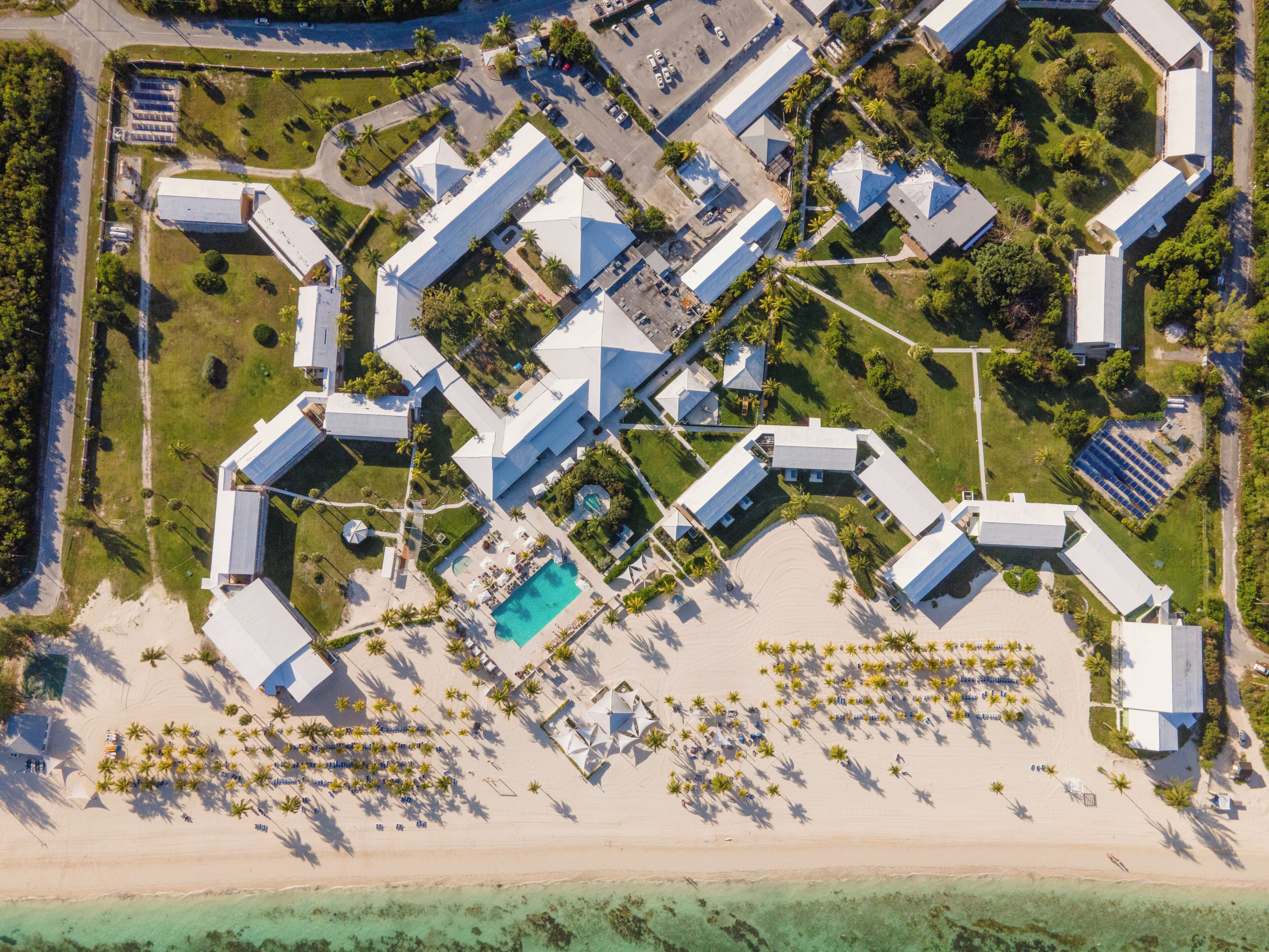 An aerial view of a resort with multiple buildings, a central pool, and a beach area with sun loungers.