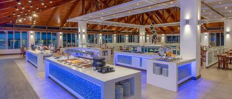 A spacious buffet area with multiple serving stations, a high ceiling with wooden beams, and large windows allowing natural light.