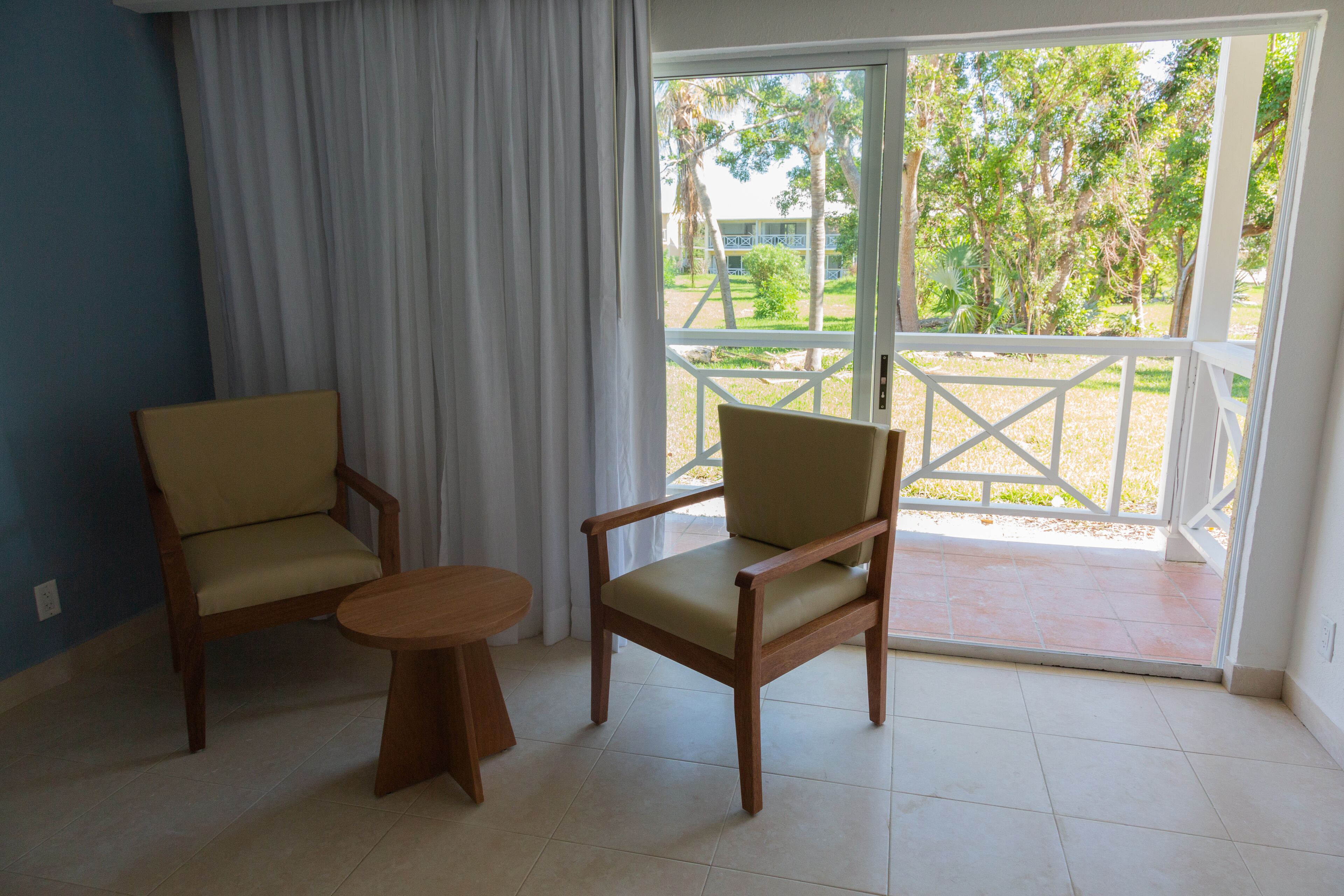 A room with two wooden chairs and a small round table, a large window with a view of greenery and a building outside.
