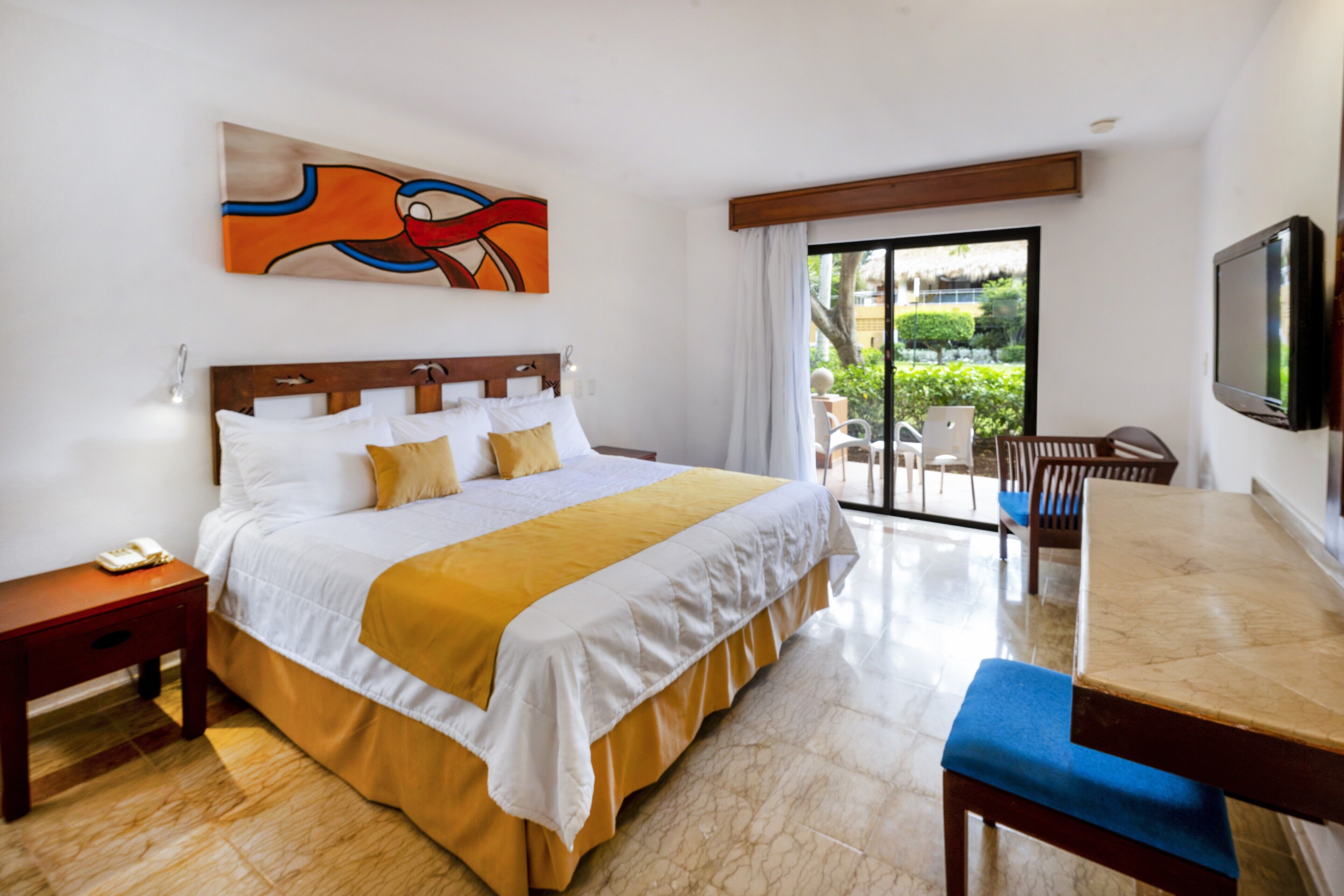A hotel room with a bed, a desk, a chair, a TV, and a balcony with a view of greenery.