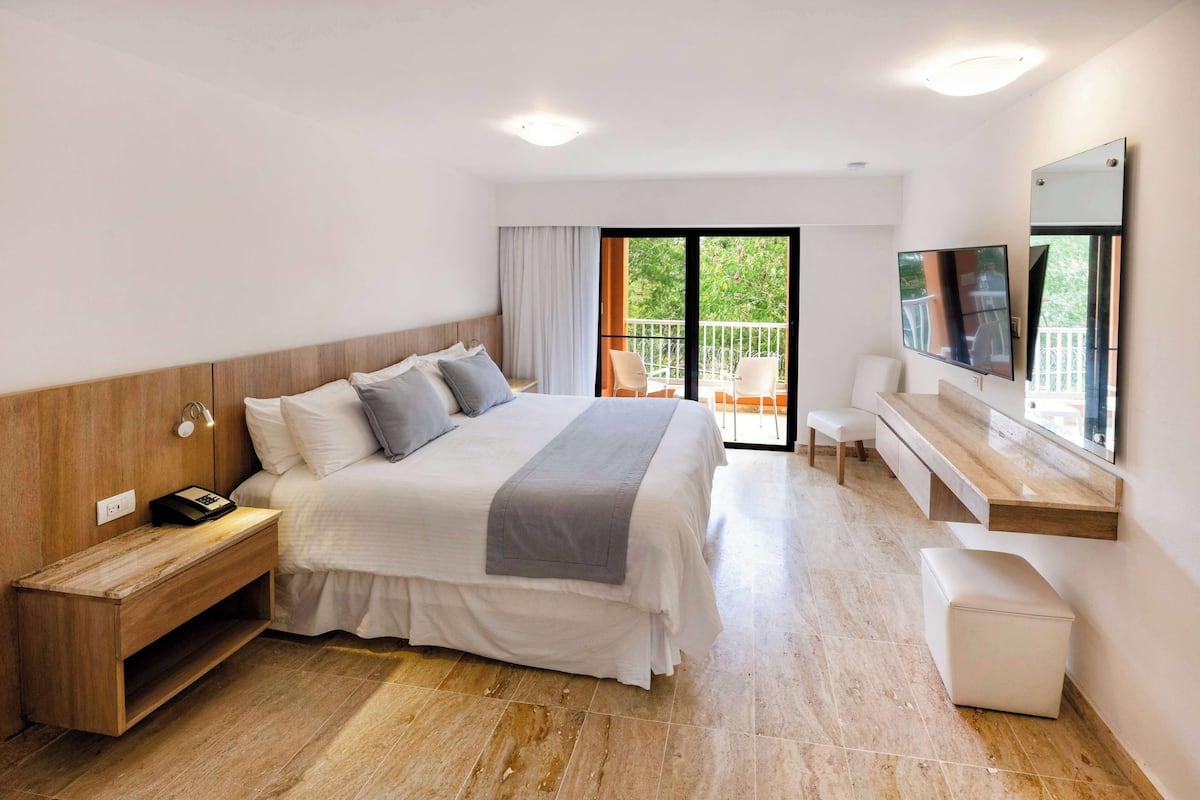 A modern hotel room with a large bed, bedside table, TV, and a balcony with outdoor seating.
