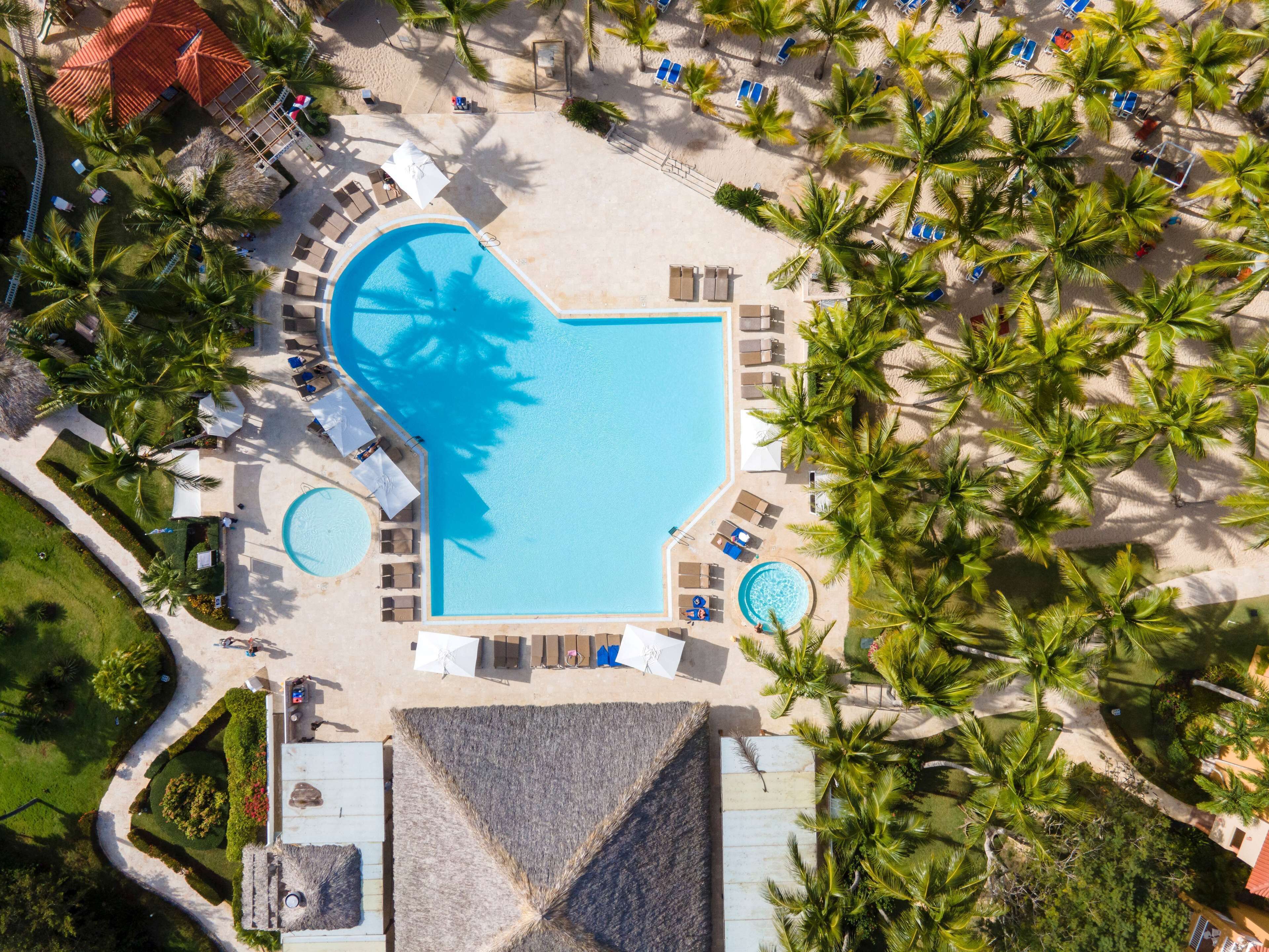 An aerial view of a resort with a large central swimming pool, multiple smaller pools, and numerous sun loungers and umbrellas.