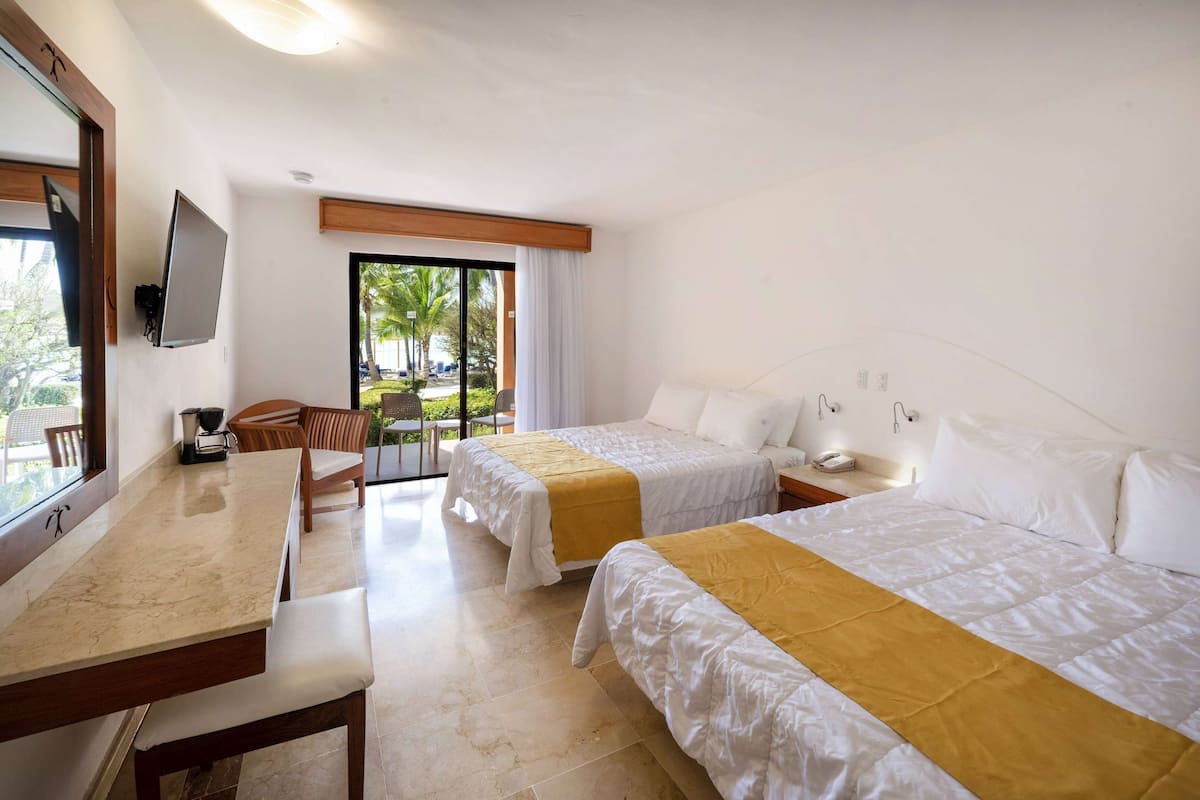 A hotel room with two beds, a desk, a chair, a TV, and a balcony with a view of palm trees.