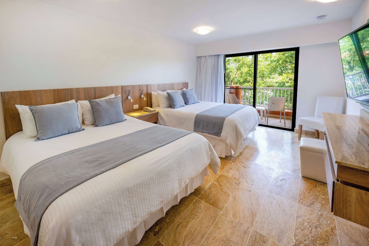 A hotel room with two beds, a desk, a chair, and a balcony with a view of trees.