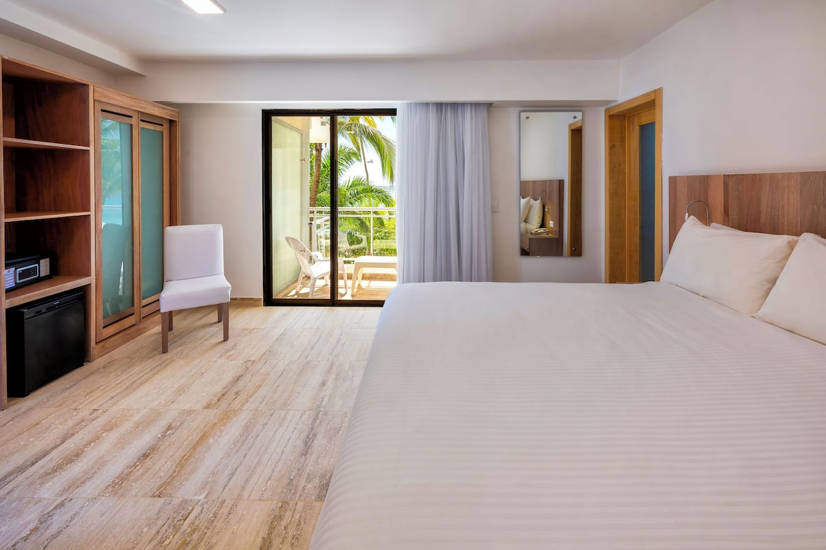 A hotel room with a large bed, a white chair, a wooden wardrobe, a small kitchen area, and a sliding glass door leading to a balcony with a view of palm trees.