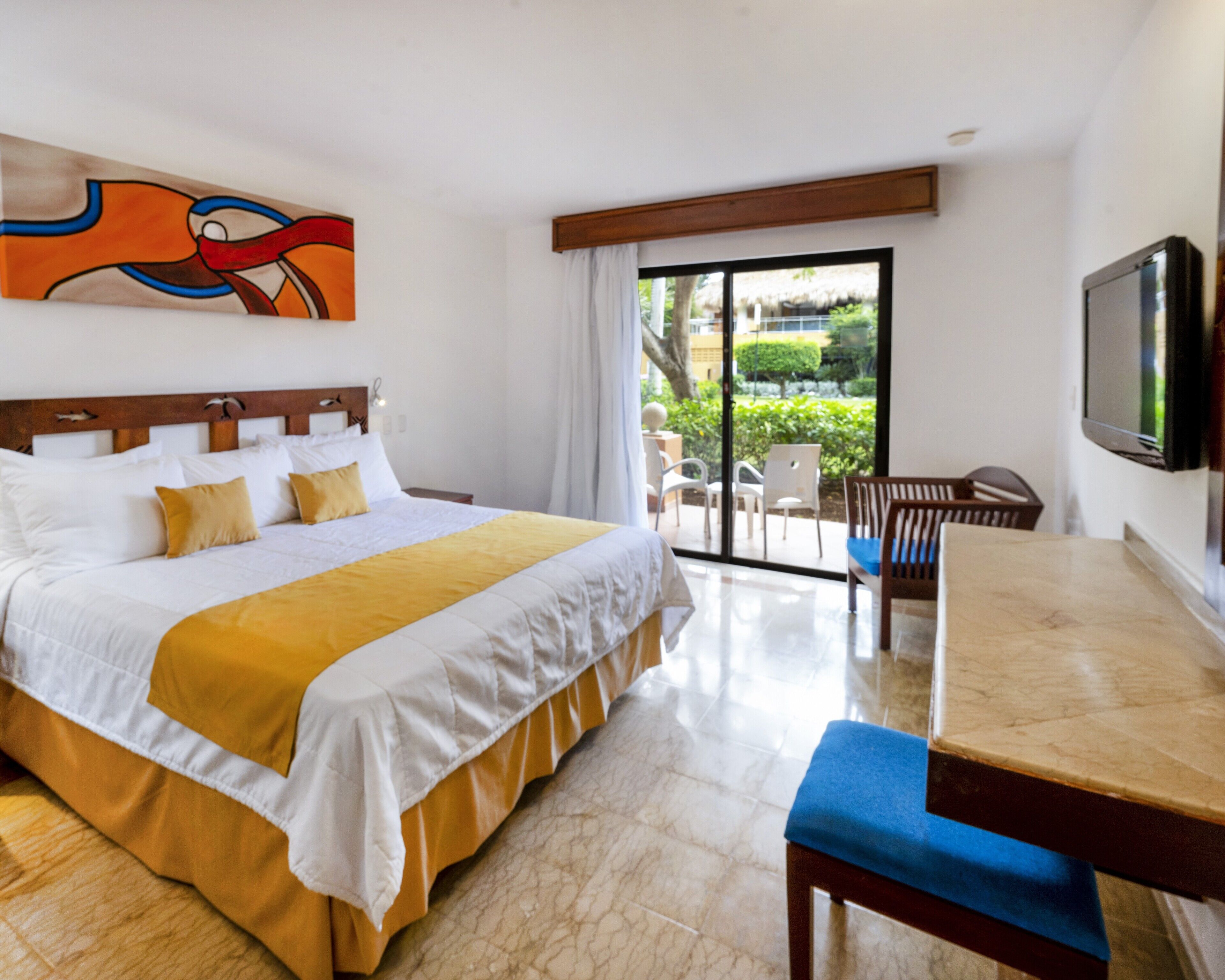 Habitación de hotel con cama, una mesa de comedor con sillas, televisor y balcón con vistas a zonas verdes.