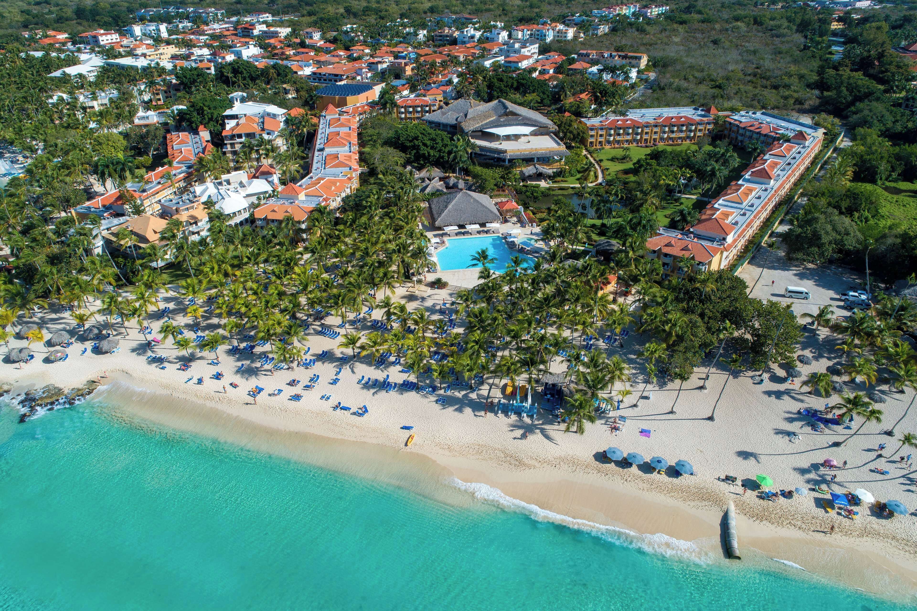 An aerial view of a resort with a beach, a swimming pool, and multiple buildings with red roofs.