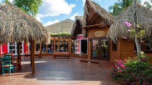 A wooden deck with thatched-roof structures, a wooden building with a sign, and tropical plants. - Viva Dominicus Palace by Wyndham, A Trademark Resort (San Rafael del Yuma)