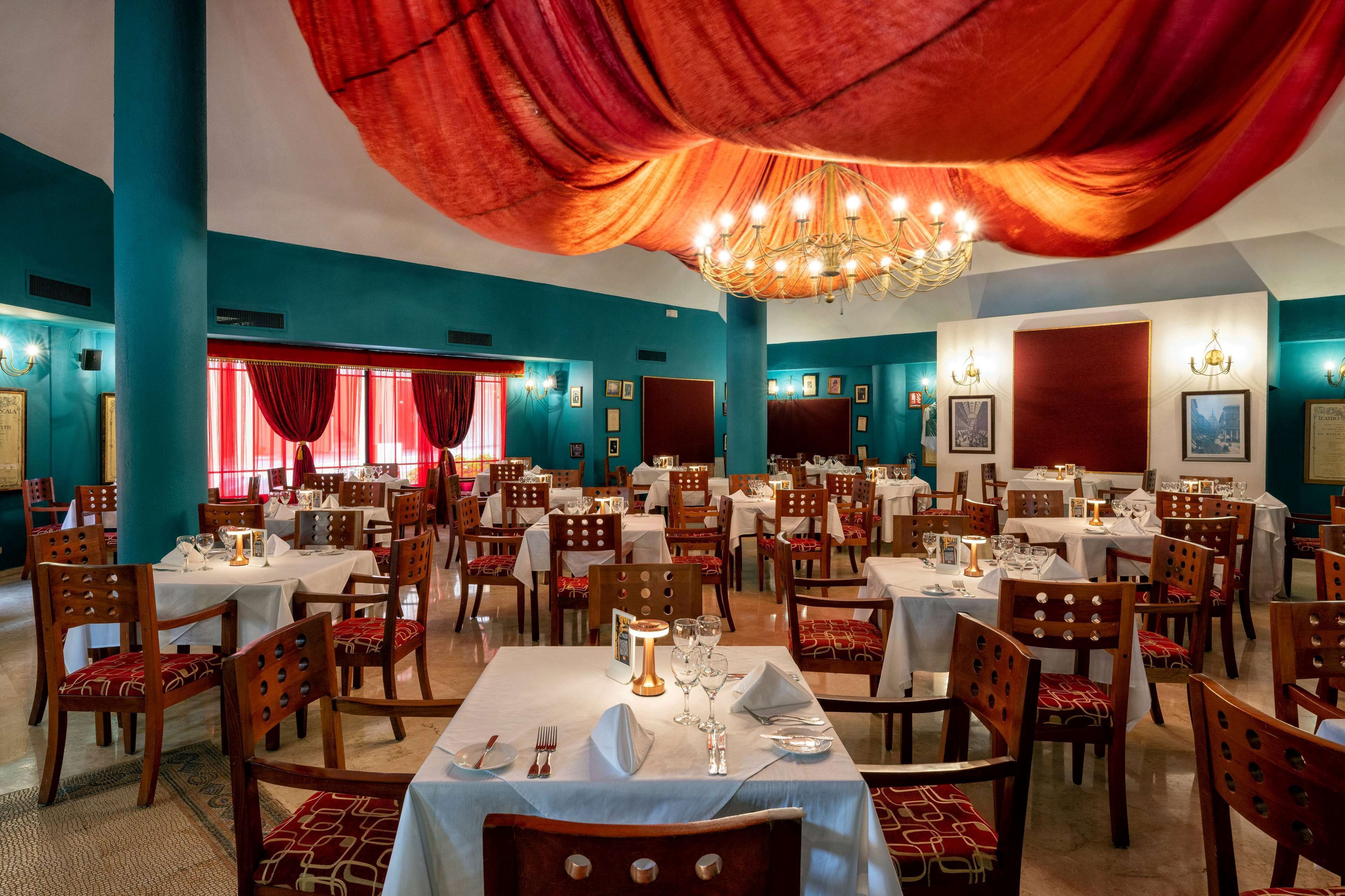 A restaurant with multiple tables and chairs, red curtains, and a chandelier.