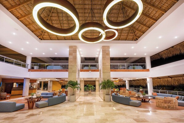 Un hall d’entrée spacieux avec un plafond haut, des luminaires circulaires et un espace de détente central agrémenté de canapés et de jardinières.