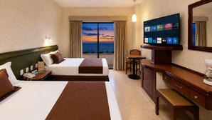 A hotel room with two beds, a TV, a desk, and a view of the ocean.