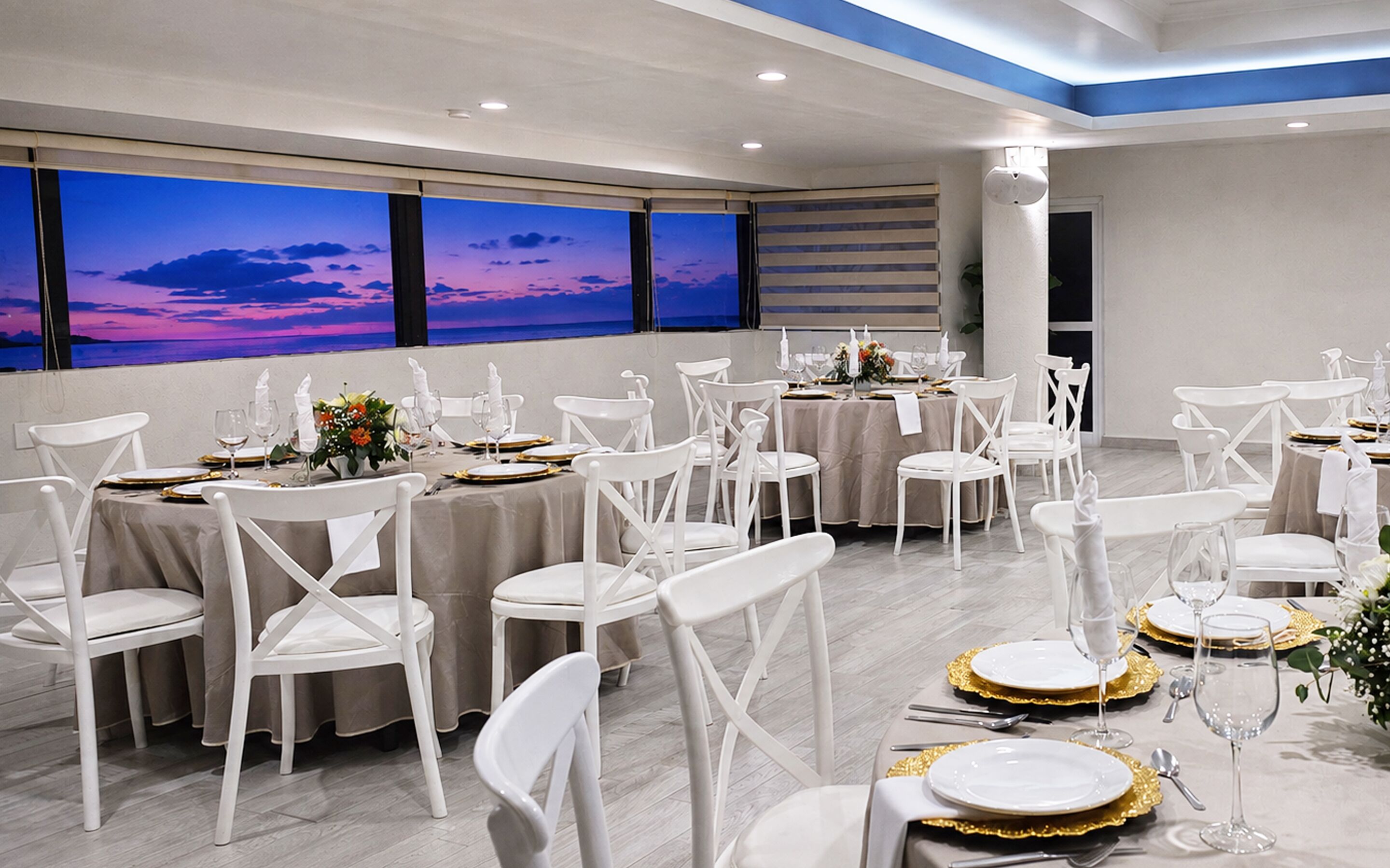 Un salón de banquetes con mesas dispuestas para un evento formal, con sillas blancas, platos con detalles dorados y vista al atardecer a través de amplias ventanas.