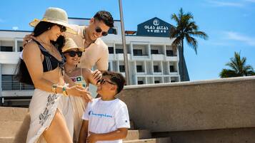 A family at a hotel with a view of the ocean and a building with 'OLAS ALEGAS' signage.