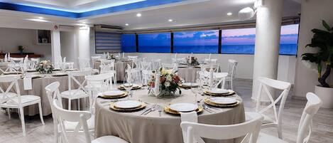 Un salón de banquetes con mesas redondas, sillas blancas y vista al océano.