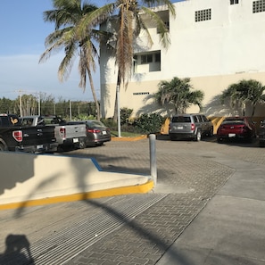 A parking area with several vehicles, including a black pickup truck and a red car, parked in front of a building with a white facade and palm trees.
