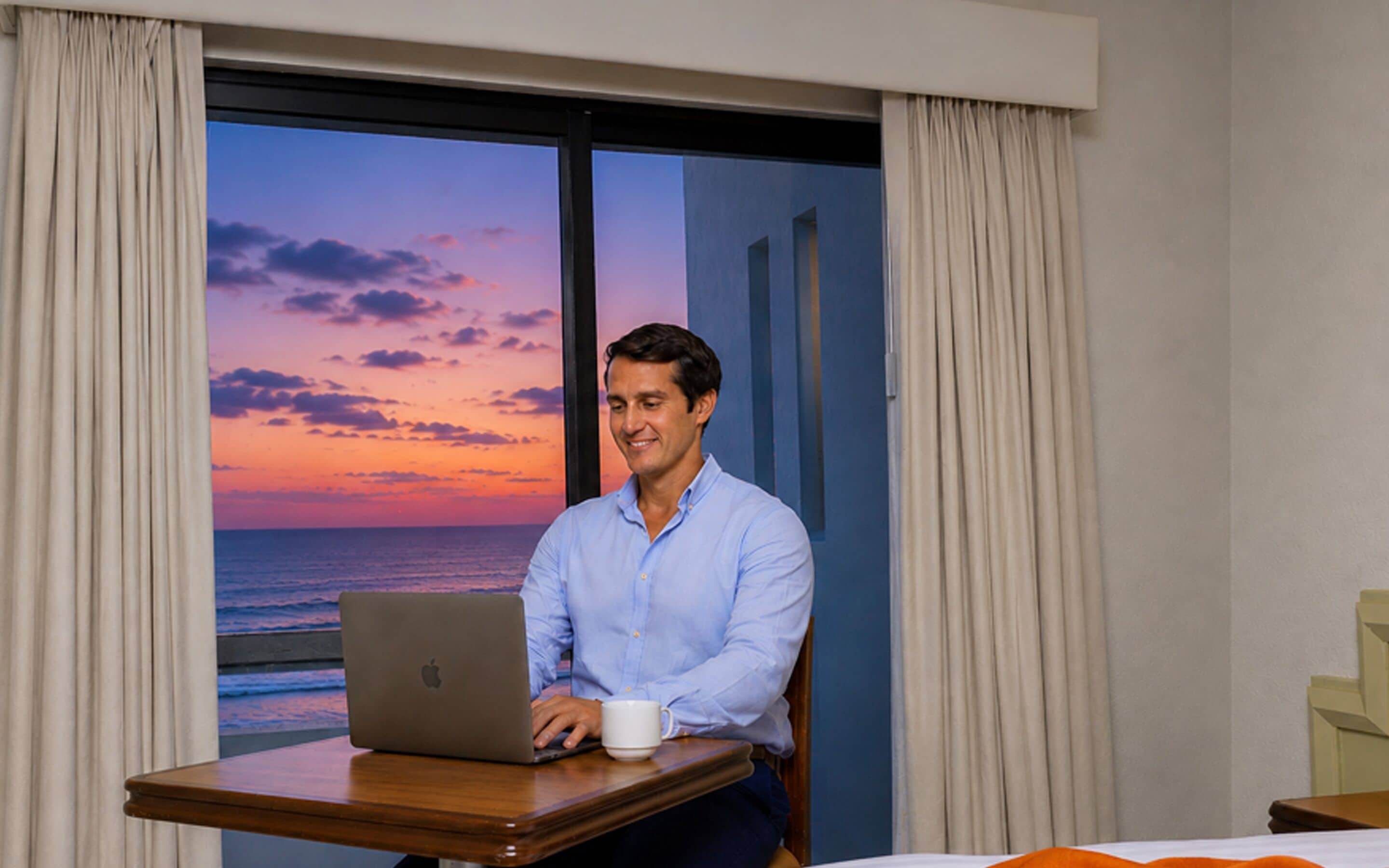 A man sitting at a wooden table with a laptop and a mug, in a room with a large window overlooking the ocean at sunset.