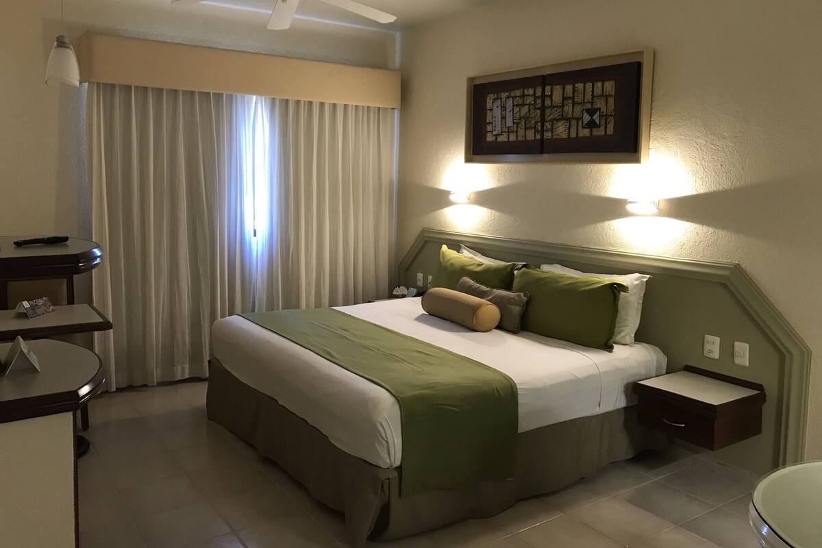 Een hotelkamer met een bed, nachtkastjes, een bureau, een plafondventilator en een raam met gordijnen.