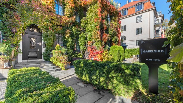 A hotel entrance with a black door, a sign reading "GABELSHUS HOME HOTEL," and a garden with neatly trimmed bushes. - Home Hotel Gabelshus (Oslo)