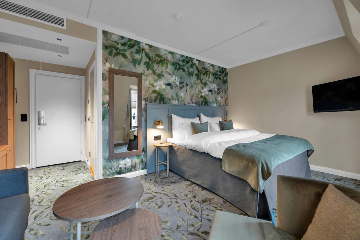 A hotel room with a bed, a small table, a chair, a mirror, and a TV on the wall.