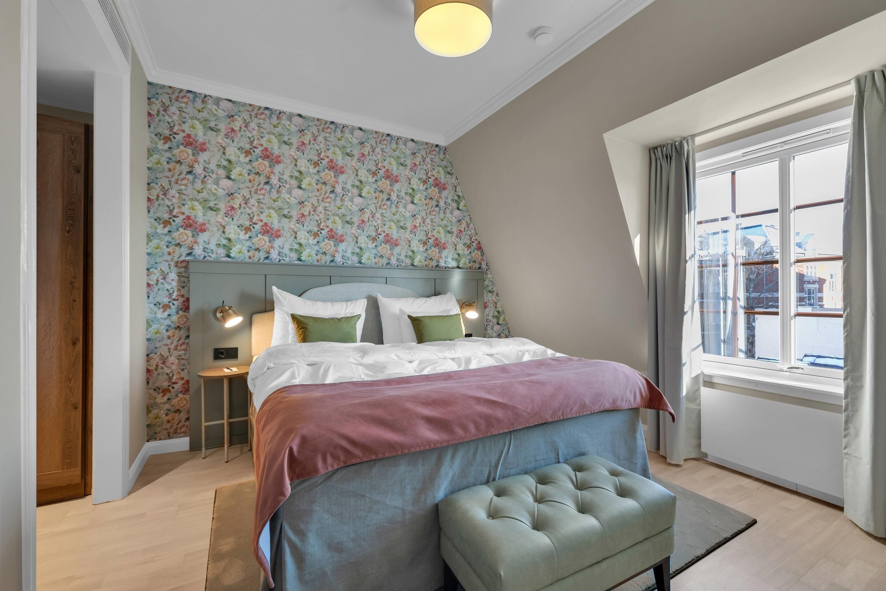 A bedroom with a bed, a bench, a nightstand, a lamp, and a window with curtains.