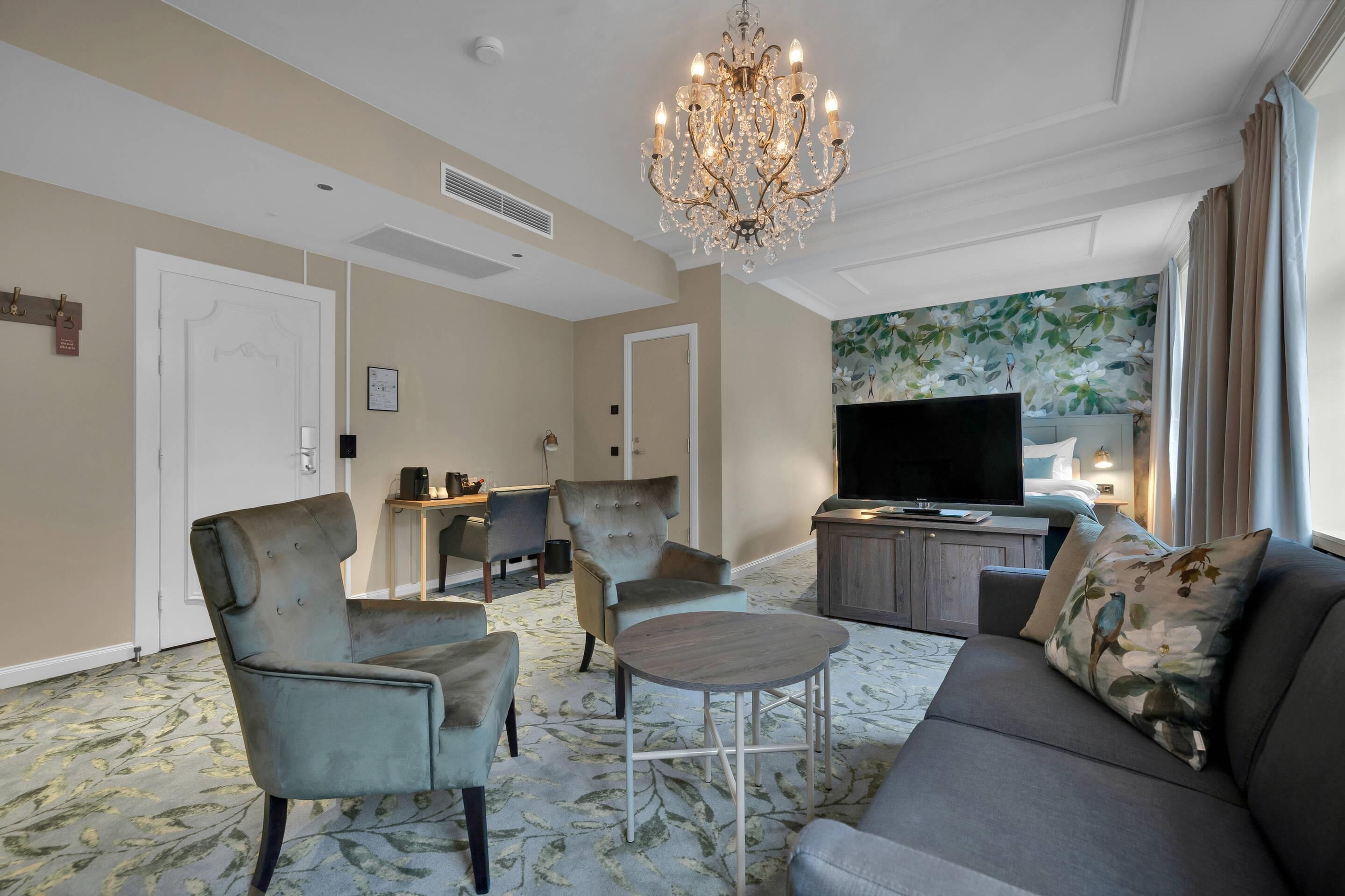 A hotel room with a sofa, armchairs, a small table, a chandelier, a TV, and a floral wallpaper.