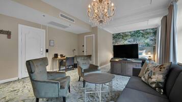 A hotel room with a sofa, armchairs, a small table, a chandelier, a TV, and a floral wallpaper.