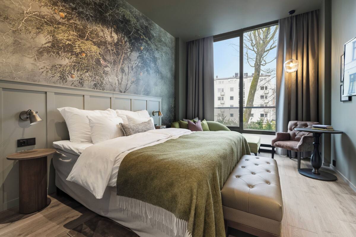 A modern hotel room with a large bed, a seating area, a view of the city, and a unique wall mural.