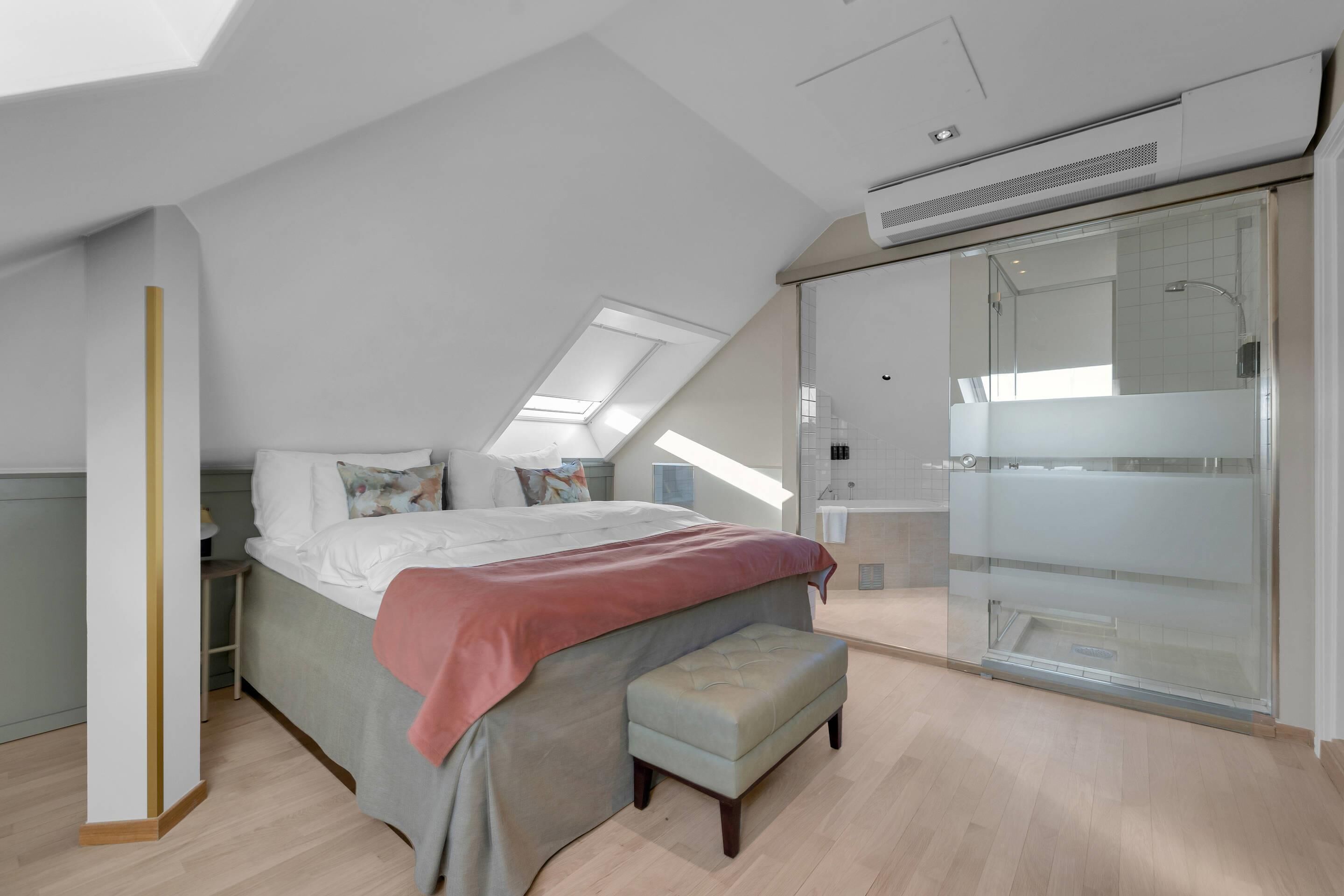 A modern bedroom with a large bed, a bench, a walk-in closet, and a skylight.