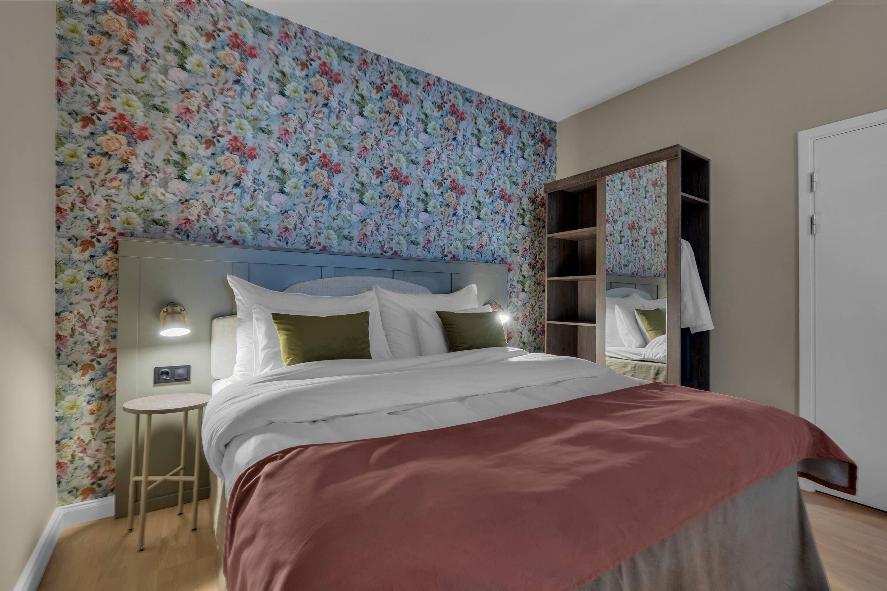 Un dormitorio con papel tapiz floral, una cama con ropa de cama blanca, una mesita de noche y un armario con espejo.