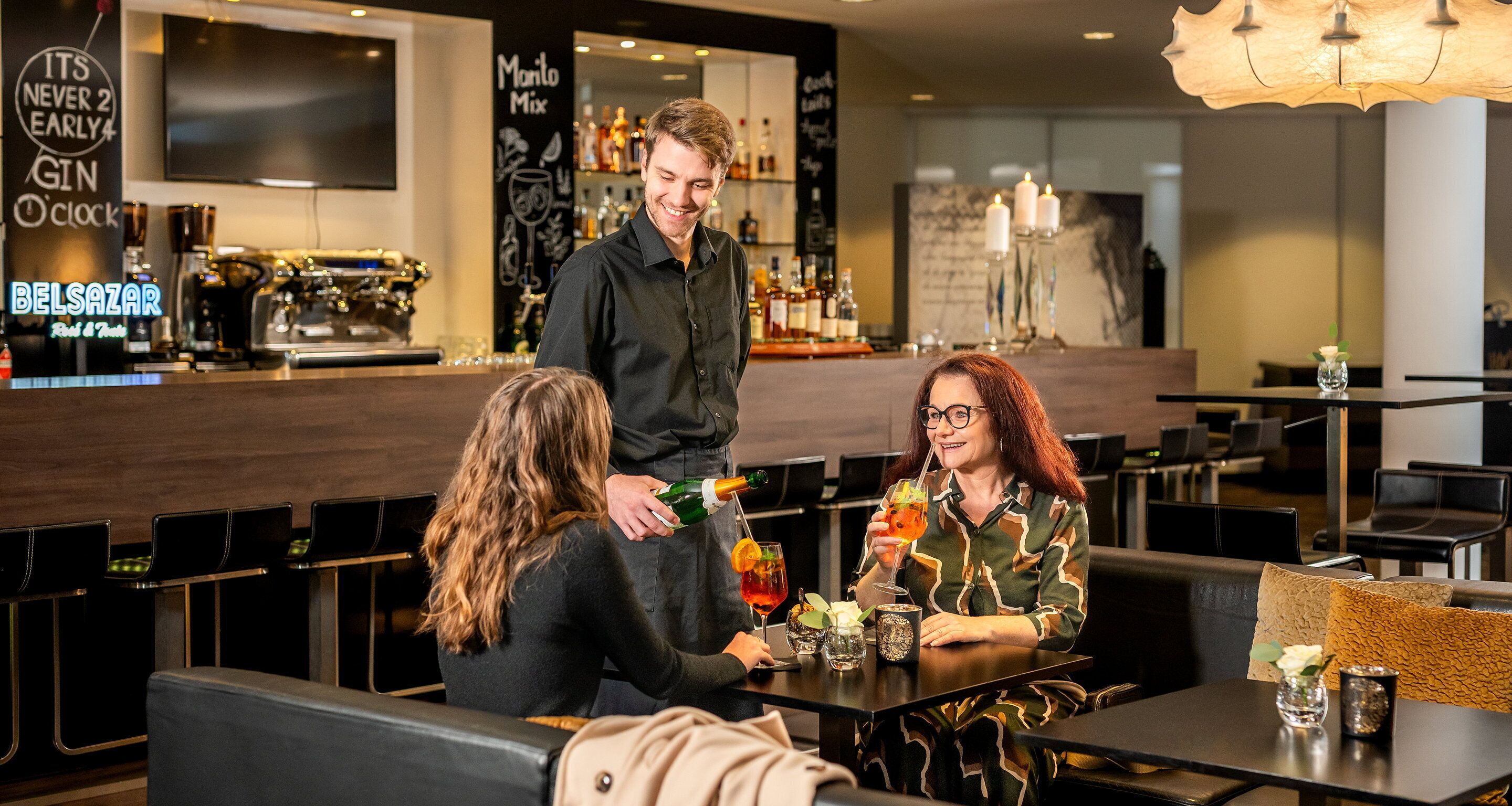 Zona de bar con un barman sirviendo bebidas a dos clientes sentados.