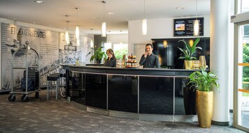 Un área de recepción moderna de hotel con un mostrador, dos empleados, un panel de menú y una planta grande en maceta.