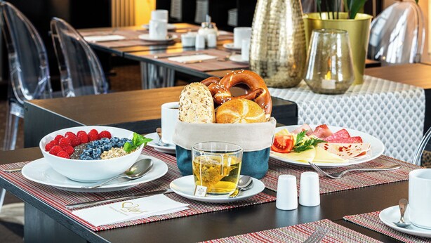 Un desayuno con un bol de bayas, una taza de té, un plato de fiambres y una cesta de pan y pretzels.