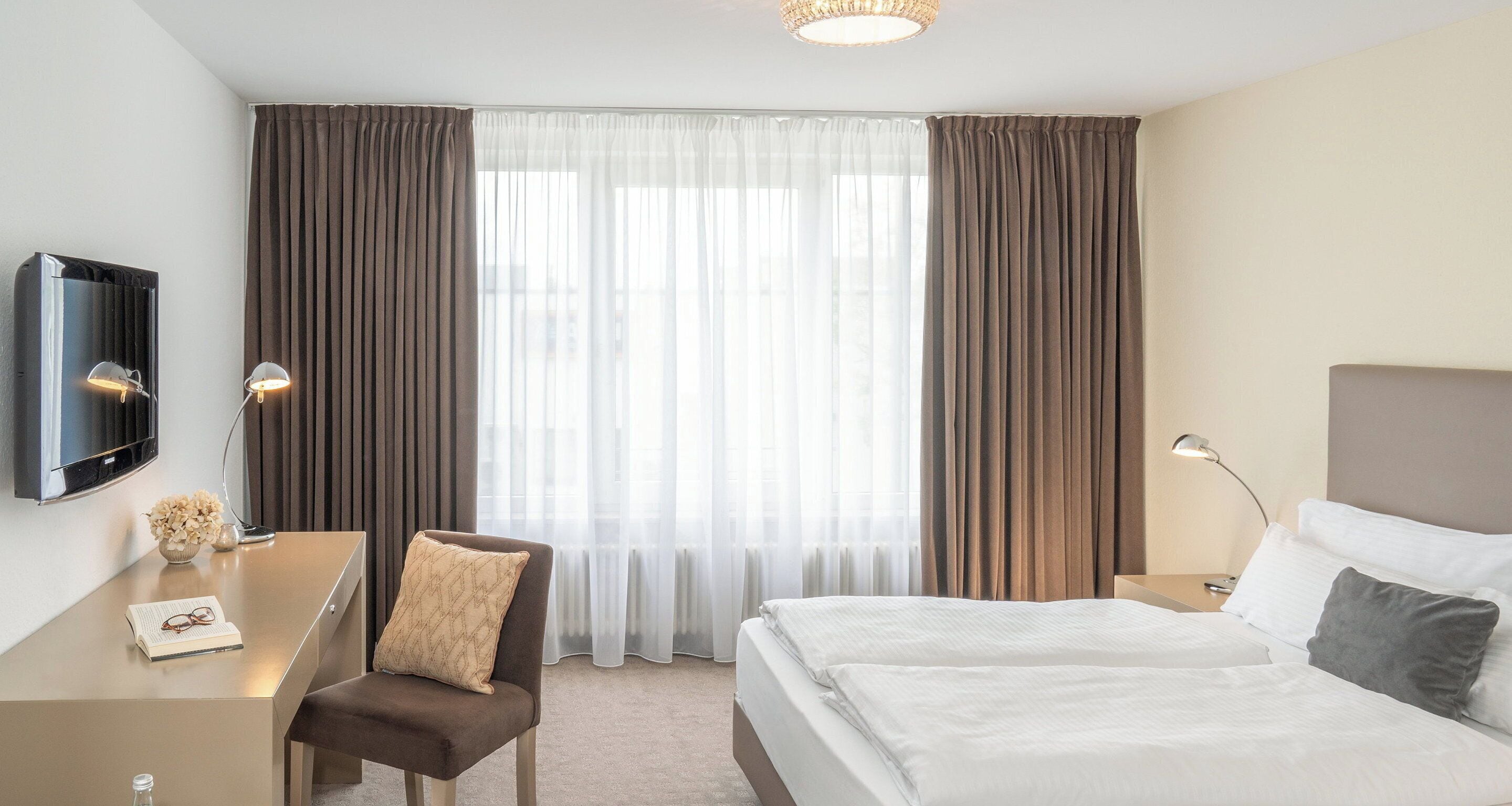 Ein Hotelzimmer mit Bett, Schreibtisch, Stuhl, Fernseher und einem Fenster mit Vorhängen.