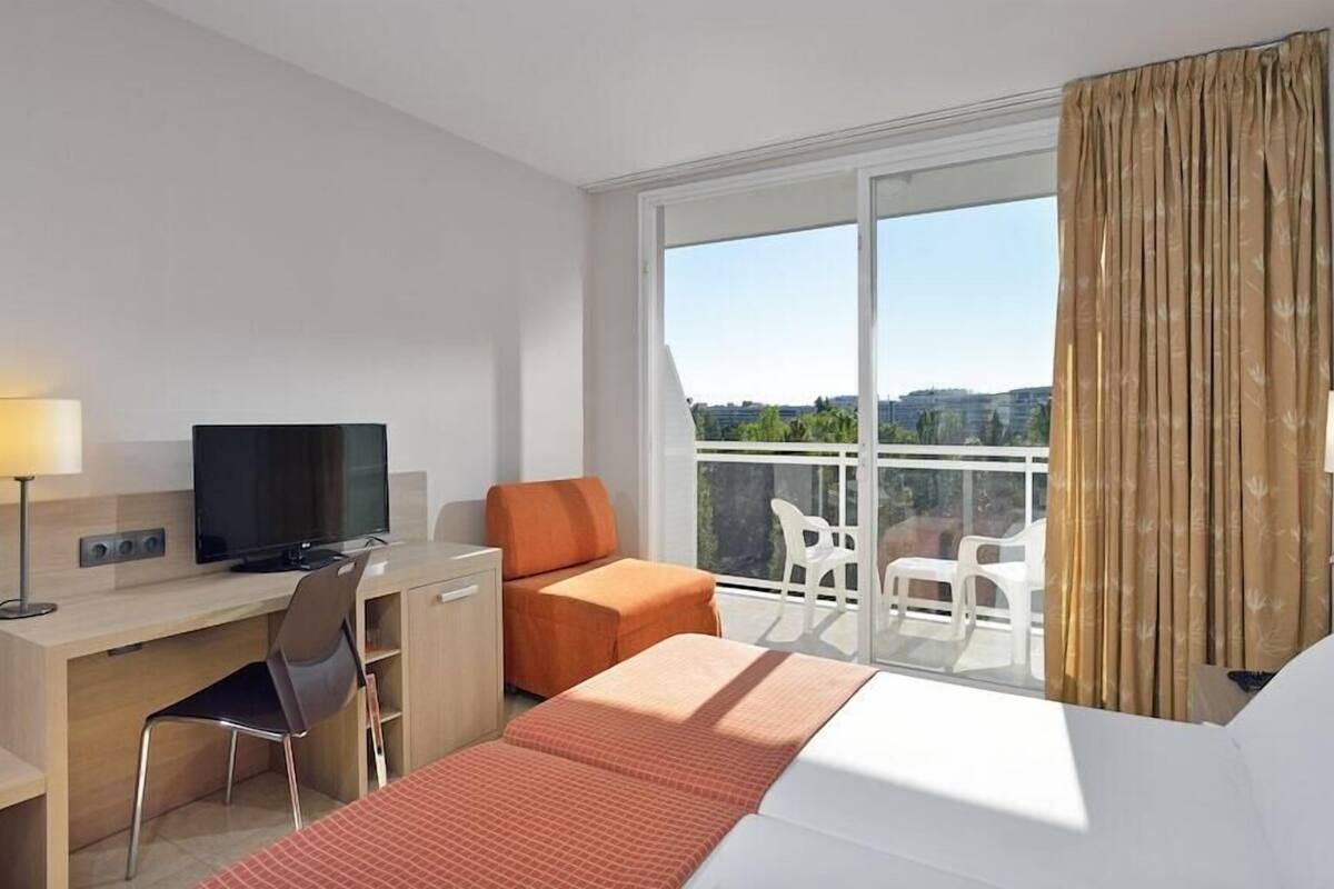 A hotel room with a bed, desk, chair, television, and a balcony with chairs.