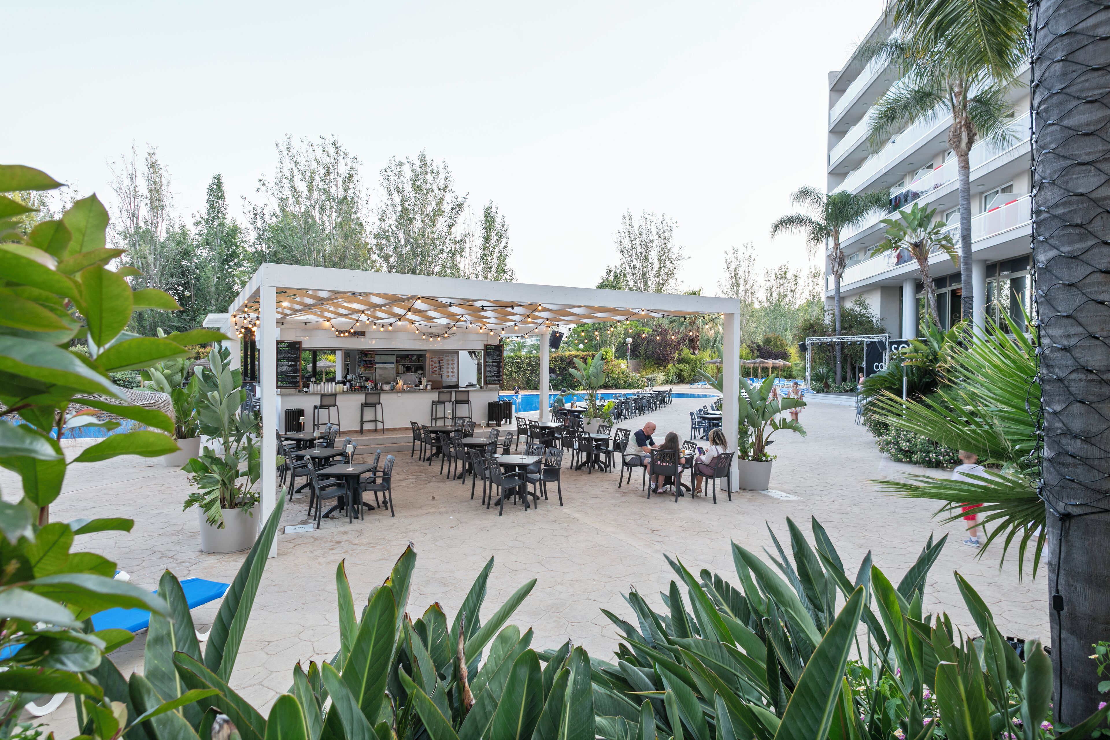 Um café ao ar livre com mesas e cadeiras, rodeado por plantas em vasos e uma piscina.