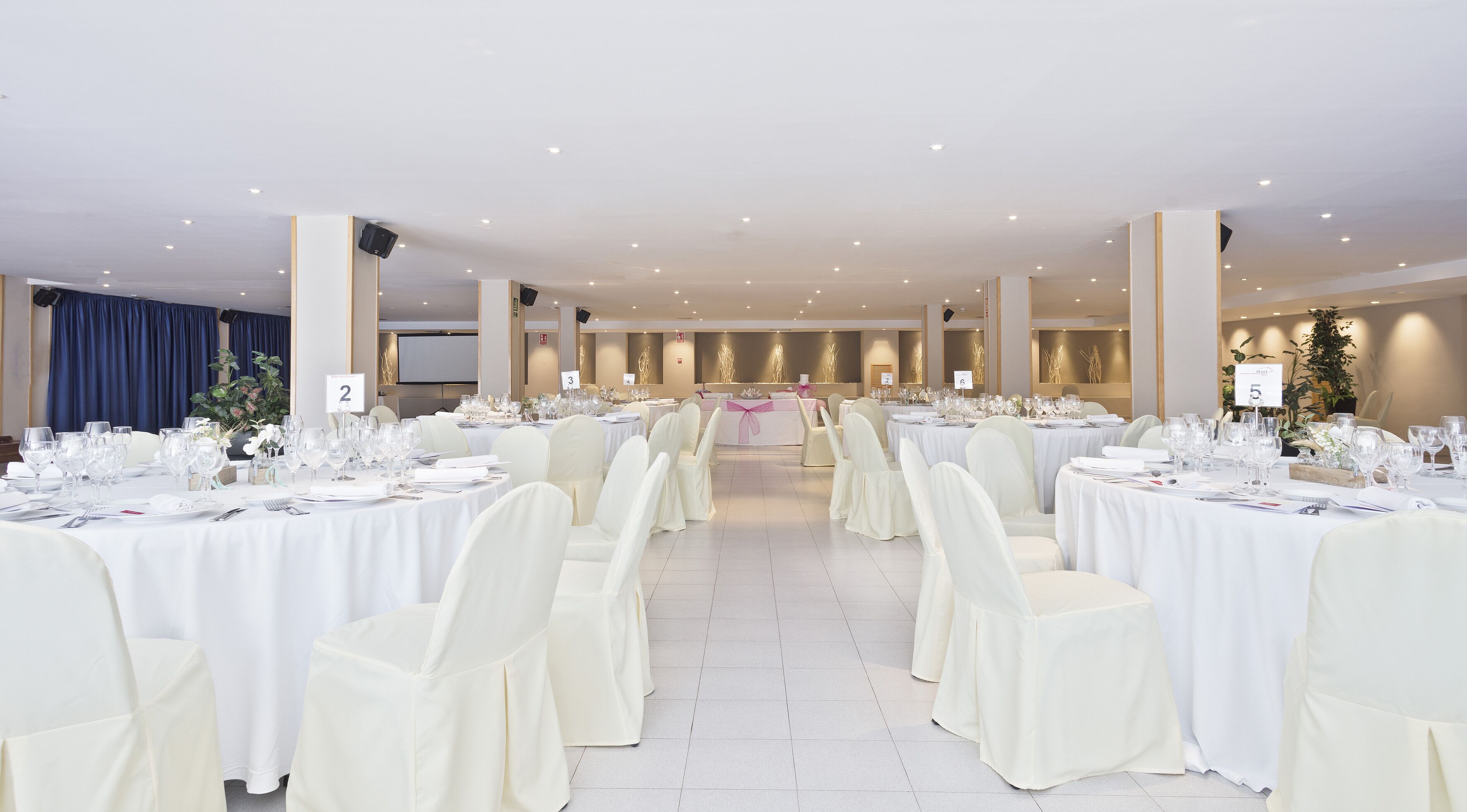 Salão de banquetes com mesas redondas, toalhas brancas e cadeiras.