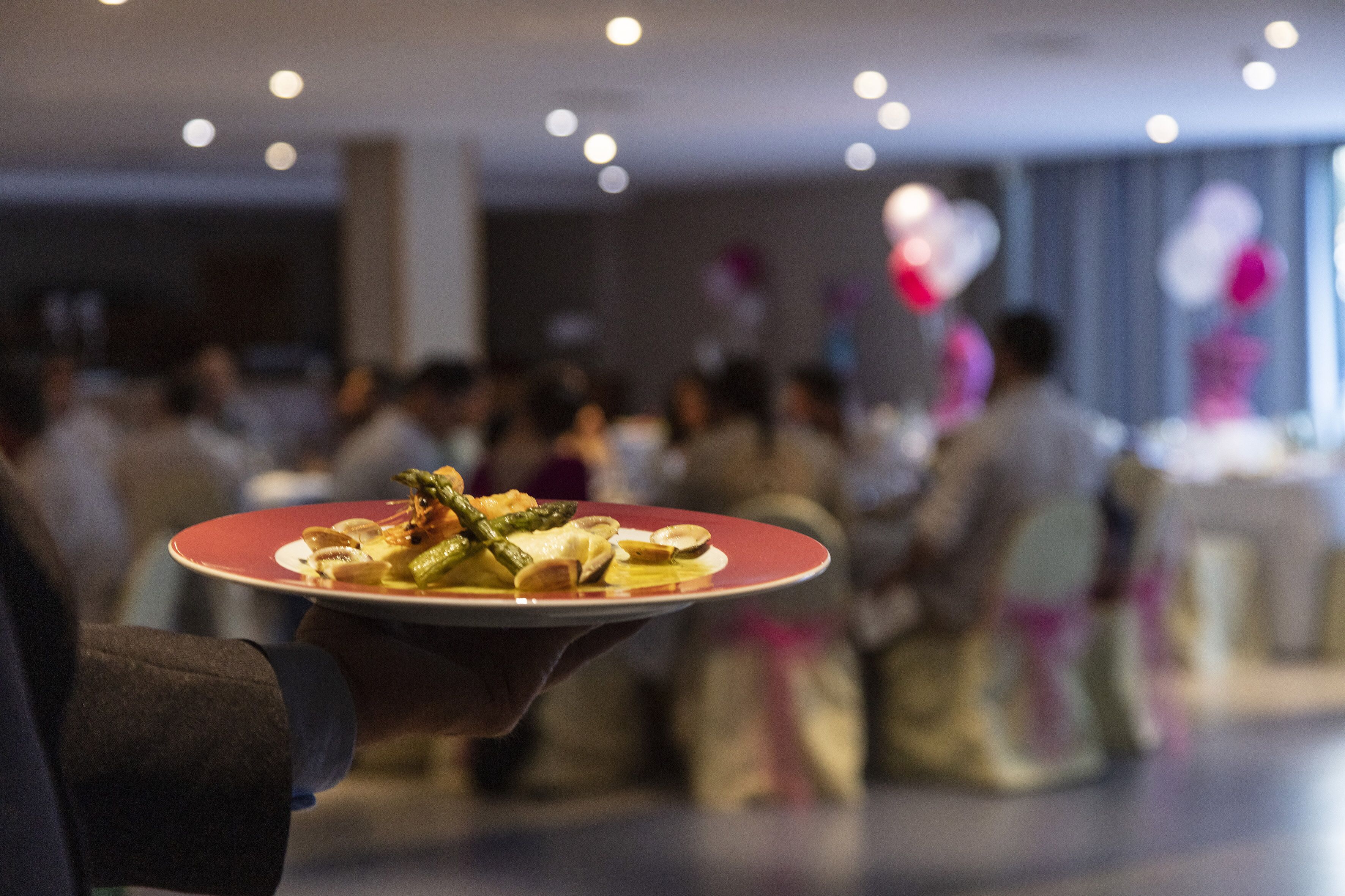 Una persona sostiene un plato con comida, con gente de fondo en una habitación.