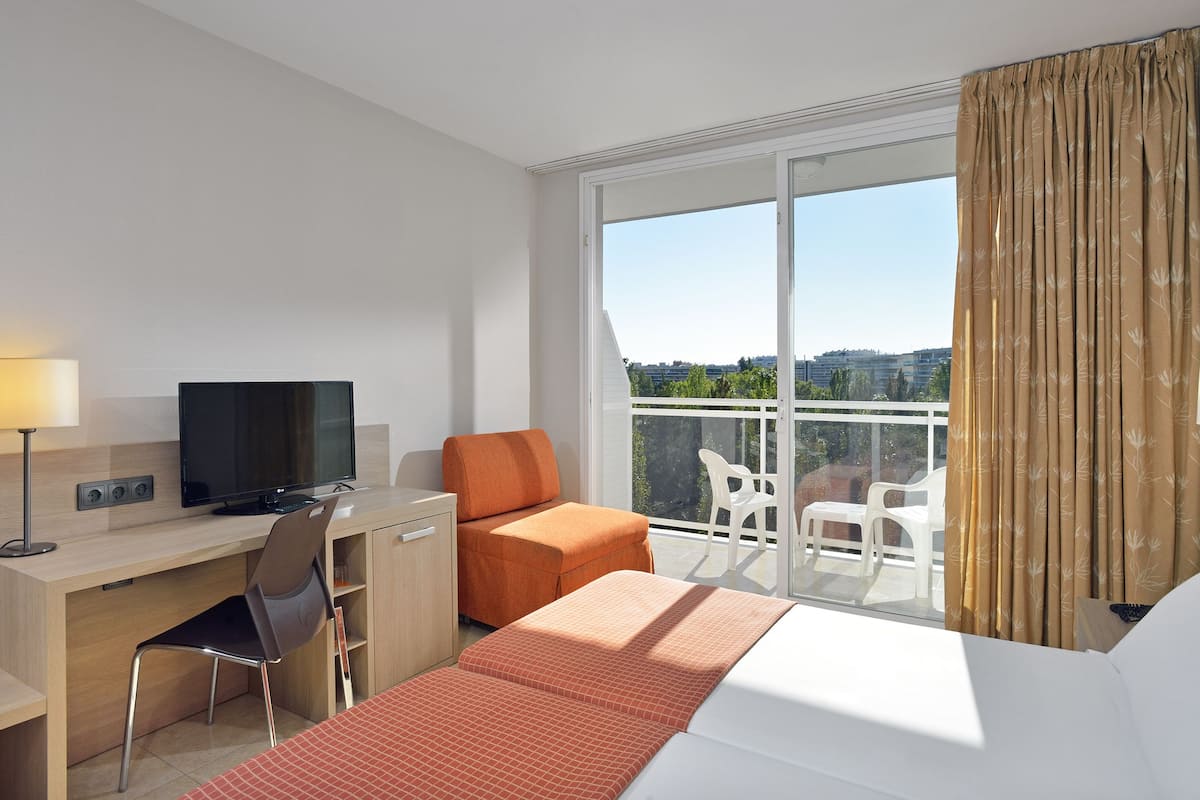 A hotel room with a bed, a desk with a computer, a TV, a chair, a balcony with a view, and a window with curtains.