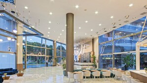 A spacious lobby with a high ceiling, large windows, and a central column. There are seating areas with tables and chairs, and a reception desk. - Sol Costa Daurada (Salou)